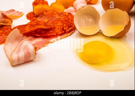 Ein zerbrochenes Hühnerei, Scheiben von Wurst und Speck in Scheiben und Tomaten auf weißem Hintergrund. Nahaufnahme Stockfoto