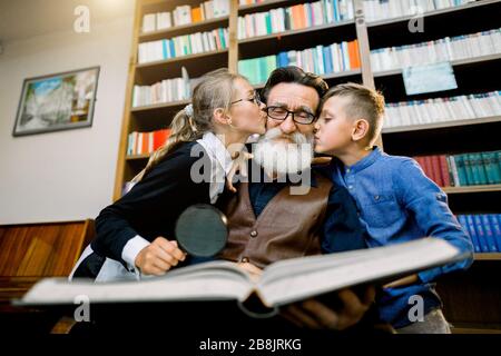 Wunderschönes Porträt glücklicher Kinder, Junge und Mädchen, die ihren alten bärtigen Opa in den Wangen küssen, während sie Zeit verbringen und zusammen erstaunliches Buch lesen Stockfoto