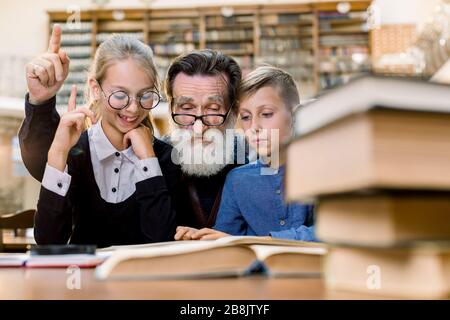 Fröhlich lächelnder Großvater liest Buch mit Enkel und Enkelin, sitzt am Tisch in alter Vintage-Bibliothek. Älterer Mann und das Mädchen sind es Stockfoto