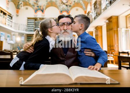 Angenehmer gutaussehender bärtiger alter Großvater, der mit seiner Enkelin und seinem Enkel Zeit in der Bibliothek verbrachte und ihn in die Wangen küsste Stockfoto