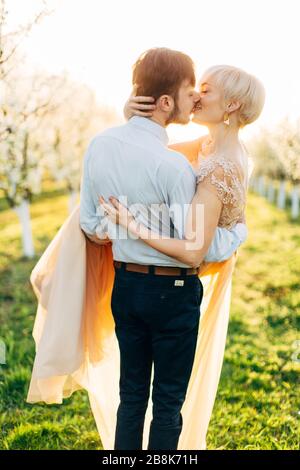 Fröhliches, küssendes Hochzeitspaar verliebt sich im schönen blühenden Garten am sonnigen Frühlingstag, Mann hält Frau an den Händen. Ein Paar im blühenden Garten zu küssen Stockfoto