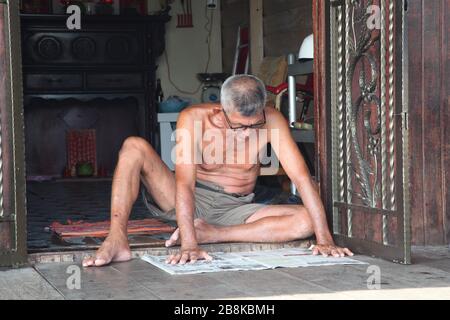 Nahaufnahme eines einheimischen Mannes in Penang, Malaysia, der auf dem Boden auf der Veranda vor seinem Haus sitzt und eine Zeitung liest Stockfoto