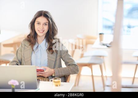 Ehrliches Porträt der hübschen jungen Frau, die während der Arbeit oder beim Lernen am Tisch in leerem Café die Kamera betrachtet und das Smartphone hält, kopiere Raum Stockfoto