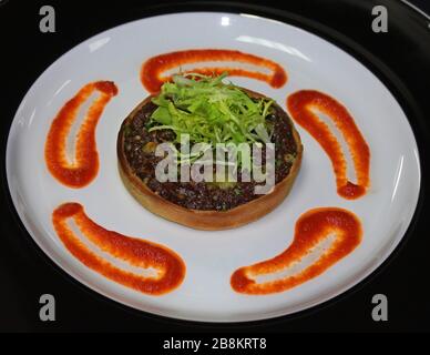 Karamellisierte Zwiebeltart mit Tomaten-Coulis und Frisee-Salat in weißer und schwarzer Platte, französische Spezialitäten. Stockfoto