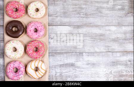 Acht traditionelle Donuts mit mehrfarbiger Glasur auf Wickelpapier auf weiß lackiertem Holzhintergrund. Donuts - traditionelles Süßgebäck. Draufsicht, Stockfoto