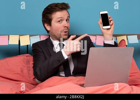 Kaukasischer reifer Mann, der im Bett liegt und sein Laptop mit dem Telefon hält und auf den Bildschirm zeigt. Bleiben Sie während der Quarantäne zu Hause. Stockfoto
