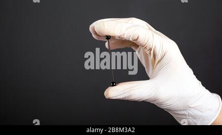 Hand in einem weißen sterilen Handschuh hält ein Piercing für Knorpelohrschmuck auf dunklem Hintergrund dicht oben. Stockfoto
