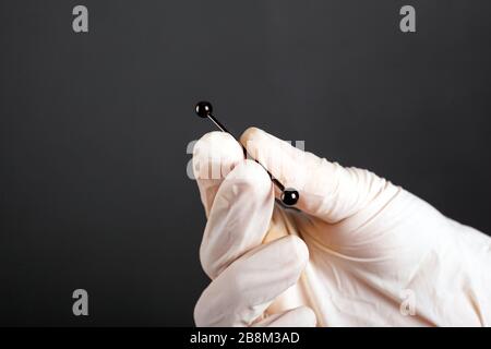Hand in einem weißen sterilen Handschuh hält ein Piercing für Knorpelohrschmuck auf dunklem Hintergrund dicht oben. Stockfoto