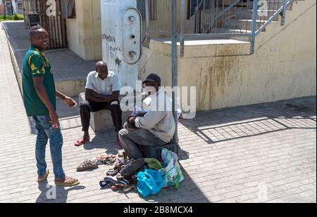 Zwelihle, Hermanus, Westkappo, Südafrika. Mann, der mit Freunden am Straßenrand die Schuhe mausiert und putzt. Stockfoto