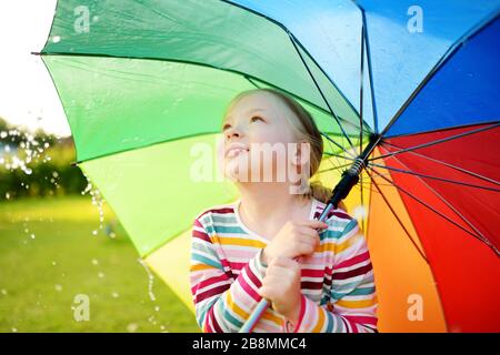 Süßes junges Mädchen mit farbenfrohem Regenbogenschirm am regnerischen Sommertag. Kinder, die unter warmem Regen im Freien laufen. Sommeraktivitäten im Freien für Kinder. Stockfoto