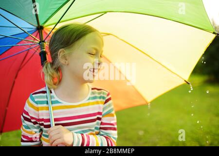 Süßes junges Mädchen mit farbenfrohem Regenbogenschirm am regnerischen Sommertag. Kinder, die unter warmem Regen im Freien laufen. Sommeraktivitäten im Freien für Kinder. Stockfoto