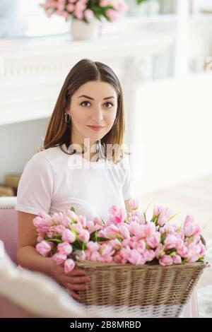 Schöne junge Frau, die auf dem Sofa sitzt und einen Korb mit rosafarbenen Tulpen hat. Stockfoto