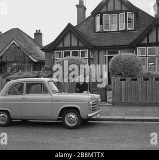 1958, historisch, auf dem Straßenbelag vor ihrer Vorstadt, einem Haus im Stil der 1930er Jahre, England, Großbritannien, eine Dame, die sich an ihr Auto lehnt, ein beliebter Motorwagen der Epoche, ein zweitüriger Ford Anglia 100E. Stockfoto