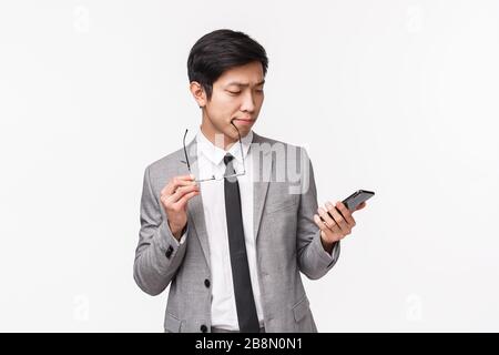 Taillen-Up-Portrait von nachdenklichen, gutaussehenden asiatischen Geschäftsleuten, die sich entscheiden, und das Handydisplay als Auswahl der Online-Bestellung ansehen Stockfoto