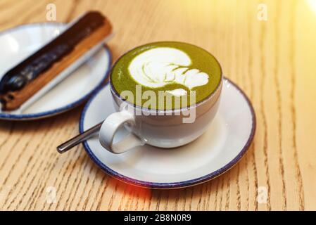 Matcha grün in einem weißen Becher auf einem Holztisch in einem Café neben einem süßen Dessert. Schokolade eclair zum Frühstück. Stockfoto