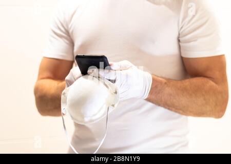 Ein Mann in einem weißen Hemd mit Schutzhandschuhen hält eine medizinische Maske und benutzt ein Handy auf weißem Hintergrund. Pandemiekonzept Stockfoto