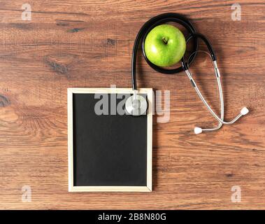 Stethoskop, Schwarzen Brett für Text und grüner apfel auf Holztisch Draufsicht. Gesunder Lebensstil, alternative Medizin oder ein Konzept für Dietenkind. Leerzeichen für Text. Stockfoto