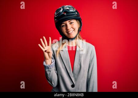 Junge, schöne blonde Motorradfahrerin, die einen Motorradhelm über rotem Hintergrund trägt und mit den Fingern Nummer vier zeigt, während sie lächelt Stockfoto