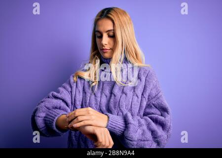 Junge, schöne blonde Frau mit legeren Turnleneck-Pullover über violettem Hintergrund Überprüfen der Zeit auf der Armbanduhr, entspannt und selbstsicher Stockfoto