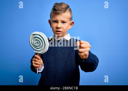 Junges kleines kaukasisches Kind, das süße Süßigkeiten-Lollipop über blauem isoliertem Hintergrund isst, ärgerte sich und frustrierte Schreie mit Wut, verrücktem und schreiendem Witz Stockfoto