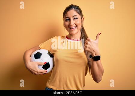 Schöne brünette Spielerin, die mit Fußball über gelbem Hintergrund Fußball spielt und mit Daumen nach oben zur Seite mit fröhlichem FA zeigt Stockfoto