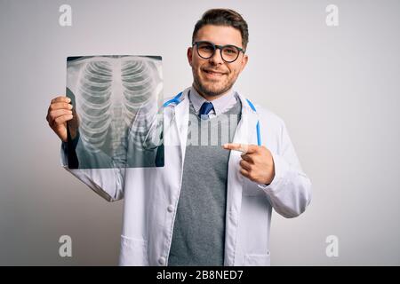 Junger Arzt, der einen medizinischen Mantel trägt, der die Röntgendiagnostik des Brustkorbs über isoliertem Hintergrund betrachtet, wobei das überraschende Gesicht mit dem Finger auf sich zeigt Stockfoto