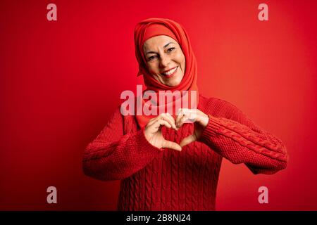Frau im mittleren Alter, die traditionellen muslimischen Hijab trägt, der über einem isolierten roten Hintergrund steht und in Liebe lächelt und Herzsymbol und Form mit den Händen zeigt. Rom Stockfoto