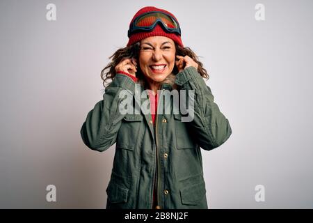 Skifahrerin im mittleren Alter, die Schneesportkleidung und eine Skibrille über weißem Hintergrund trägt, die Ohren mit Fingern bedeckt und sich ärgert über den Lärm ausdrückt Stockfoto