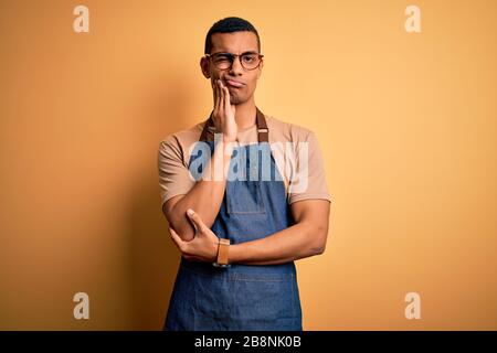 Junger, gutaussehender afroamerikanischer Ladenbesitzer, der Schürze über gelbem Hintergrund trägt und dabei müde und gelangweilt mit Depressionsproblemen mit cro aussieht Stockfoto