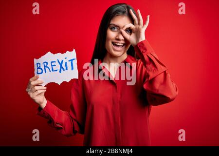 Junge schöne Frau mit blauen Augen, die Papier mit brexit Nachricht über rotem Hintergrund hält, mit fröhlichem Gesicht lächelnd, mit einem guten Schild mit Hand-auf-Augen-Blick Stockfoto