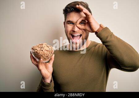 Junger blonder Mann mit Bart und blauen Augen, die Schüssel mit Cornflakes halten, mit fröhlichem Gesicht lächelnd, ok, mit Hand auf Auge, der durch f blickt Stockfoto