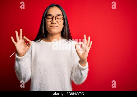 Junge, schöne asiatische Frau mit legeren Pullover und Brille über rotem Hintergrund entspannt und lächelnd mit geschlossenen Augen, die mit Meditationsgeste tun Stockfoto