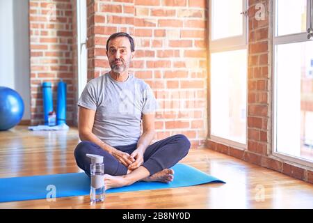 Mittelaltersportler, der auf der Matte sitzt und in der Sporthalle ausdehnende Yoga-Übungen mit lustigen Gesichtern macht. Mund aufgeblasen mit Luft, verrückt vor Stockfoto
