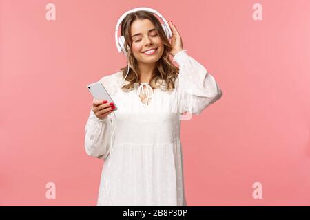Porträt eines zärtlichen, fröhlichen und süßen blonden Mädchens in weißem Kleid, mit geschlossenen Augen und einem Lächeln, das sich wie Musik in Kopfhörern anhört, Wärme fühlt und Stockfoto