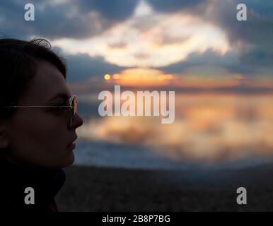 Peinlich düsteres, melancholes Frauenporträt im Profil in goldener runder Sonnenbrille mit geschälten Haaren gegen den sehr schönen Sonnenuntergang am Meer in der Abenddämmerung. Natur Stockfoto