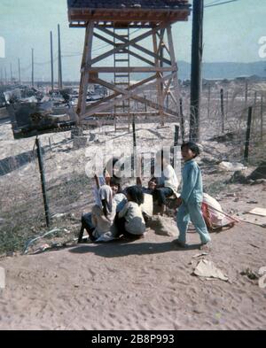 1968, Danang, Vietnam-Gebiet Vietnamesische Kinder, die während des Krieges im Danang-Flughafenbereich mit einem Wachturm und Sandsack im Hintergrund herumlungern. Stockfoto