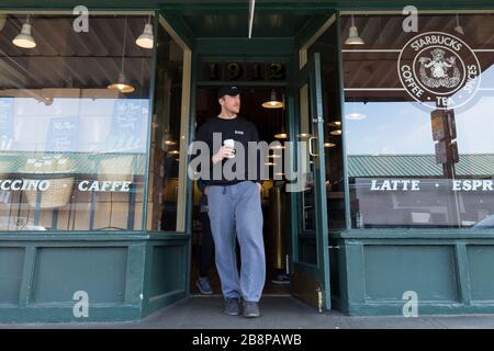 Ein Kunde verlässt am Sonntag, 22. März 2020, den ursprünglichen Starbucks am Pike Place Market mit einem Kaffee. Um die Verbreitung des COVID-19-Virus zu begrenzen, beschränkt das Unternehmen die Anzahl der Kunden, die den normalerweise überfüllten Laden gleichzeitig betreten. Stockfoto