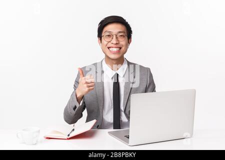 Porträt des fröhlichen und fröhlichen jungen asiatischen männlichen Unternehmers, Büromanager, der an einem Projekt arbeitet, Sitztisch mit Notizbuch und Laptop, Anrufe entgegennehmen Stockfoto