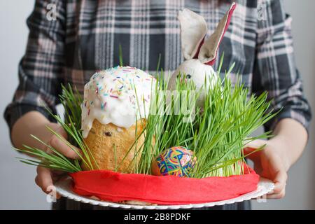 Traditioneller Osterkuchen, Eier und Häschen im Gras in Frauenhand. Osterbrot mit weißer Creme und farbenfroher Dekoration auf der Oberseite. Feasting Stockfoto