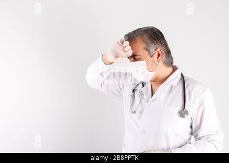Leitender männlicher Arzt, der Maske und Handschuhe trägt und die Stirn berührt, wobei die Hand im Krankenhaus gegen die weiße Wand steht Stockfoto