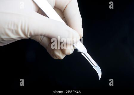 Die gerillte Hand des Chirurgen hält gebogene Skalpelle der Klinge auf schwarzem Grund. Stockfoto