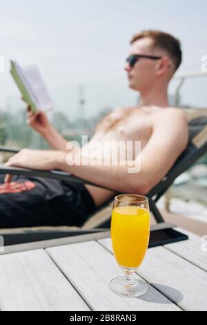 Ein Glas kalter, erfrischender Orangensaft auf dem Tisch neben dem jungen Mann, der sich in der Chaiselounge entspannen und ein Buch lesen kann Stockfoto