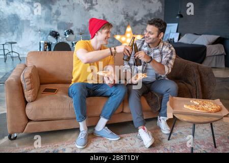 Zwei junge Männer sitzen auf dem Sofa und genießen Pizza und Bier Stockfoto