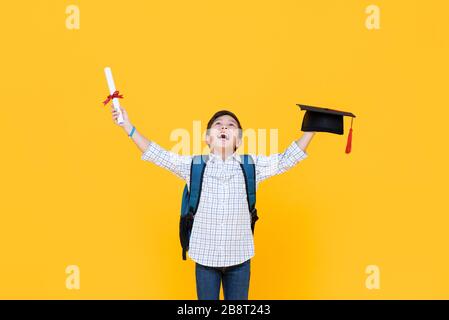 Glücklicher Absolvent Junge mit akademischer Kappe lächelnd und die Hände heben, die den Schulungstag feiern, isoliert auf gelbem Hintergrund Stockfoto