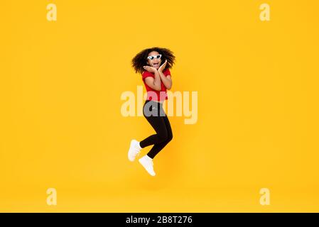 Fröhliches, modisches afroamerikanisches Mädchen, das mitten in der Luft springt und mit Händen am Kinn isoliert auf gelbem Hintergrund liegt Stockfoto