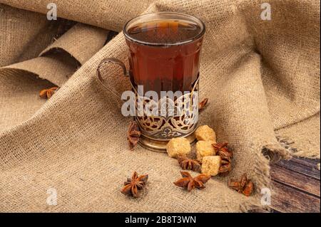 Ein facettiertes Glas Tee in einem Vintage-Cup-Halter, braunem Rohrzucker und Schokolade auf dem Hintergrund grober Homespun-Qualität. Nahaufnahme Stockfoto