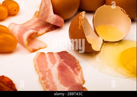 Ein zerbrochenes Hühnerei und Scheiben von Speck und Wurst und Tomaten auf weißem Hintergrund. Nahaufnahme Stockfoto