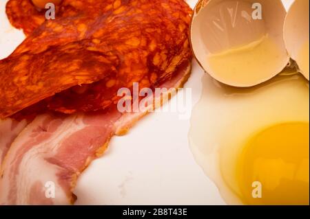 Ein zerbrochenes Hühnerei und Scheiben von Speck und Wurst auf weißem Hintergrund. Nahaufnahme Stockfoto