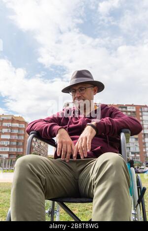 Kaukasischer Mann mit Brille und Hut depressiv und traurig in einem einsamen Rollstuhl in einem Park mit Himmel und einigen Gebäuden im Hintergrund mit Raum Stockfoto