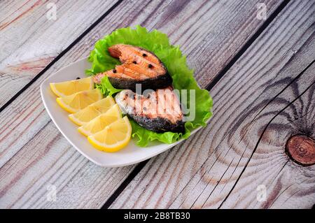 Zwei gegrillte rosafarbene Lachssteaks auf einem Salatblatt liegen auf einem Teller mit Zitronenscheiben auf einem Holztisch. Nahaufnahme. Stockfoto